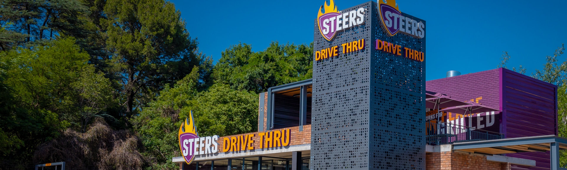 Steers Drive Thru restaurant exterior with branded signage tower surrounded by trees on a sunny day Steers Drive Thru restaurant exterior with branded signage tower surrounded by trees on a sunny day