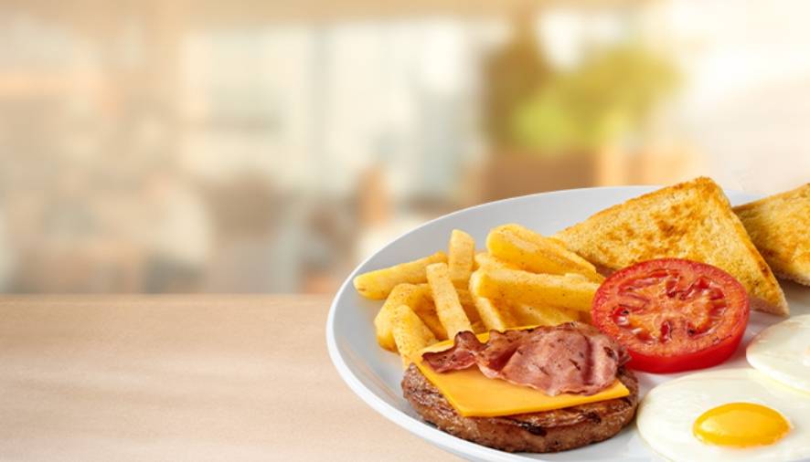 Sit-down breakfast plate with fried eggs, beef patty topped with bacon and cheese, chips, grilled tomato and toast