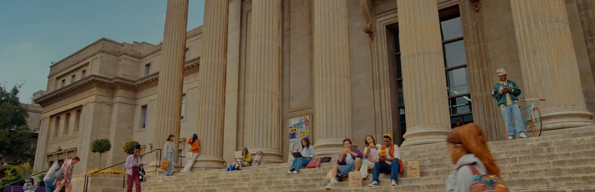 Students relaxing on stone steps outside a grand university building with tall classical columns on a sunny day