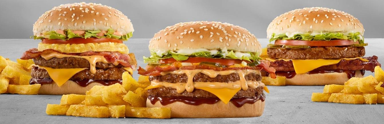 Three loaded bacon cheeseburgers with melted cheddar cheese, creamy sauce, lettuce, tomato, sesame seed buns, barbecue sauce, and chips